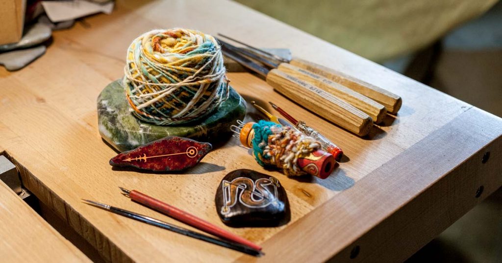 On voit différents outils des Artisans qui se sont retrouvés pour fêter Lughia : pelote de laine filée main, tricotin, pierre peinte, ciseaux à bois, tournevis électrique, plumes à dessin...